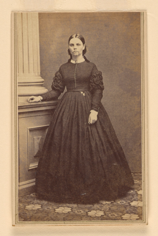 A full-length photo of Olive Oatman standing in a floor-length, long-sleeved dark dress. She is posing with one hand resting on a decorative wall. She looks at the camera but isn't smiling. Her face tattoo is visible even from a distance.