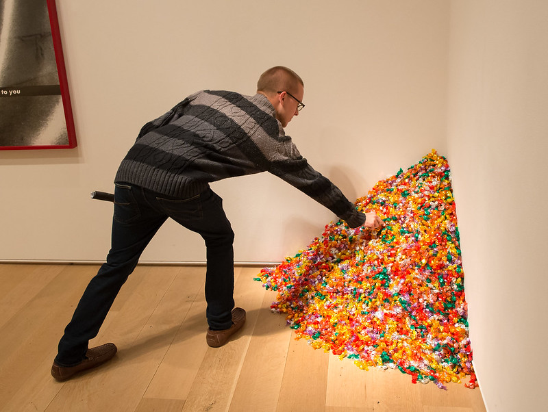 Photo shows a museum exhibit of colorfully wrapped candy piled into a heap in the corner of a room. A museum patron bends down to pick a piece of candy from the pile.