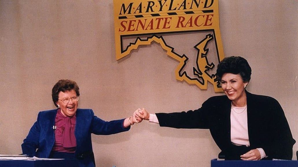 Photo of Barbara Mikulski and Linda Chavez at the 1986 Maryland Senate Race debate. Both women are smiling and appear to by laughing as the clasp each other's hands in a gesture of support and triumph.