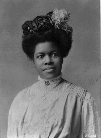 Black and white photograph of Nannie Helen Burroughs circa early 1900s. Burroughs is facing the camera but not looking directly at it. She is wearing a long sleeved dress, heart-shaped necklace, and floral headpiece.