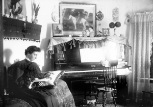 A seated woman holds a pillow and a book on her lap as she poses to the side of a piano