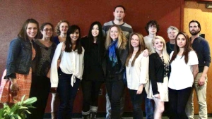 a group of thirteen people standing together and smiling in front of a dark red wall. They are in two rows: six in the front and seven in the back. Eileen Schell is the third person from the left in the front row. Minnie Bruce Pratt is the second person from the right in the back row